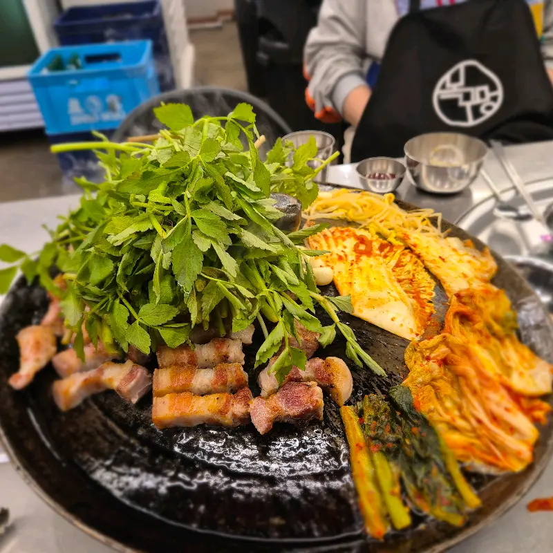 솥뚜껑에 푸짐하게 올려진 삼겹살, 미나리, 김치