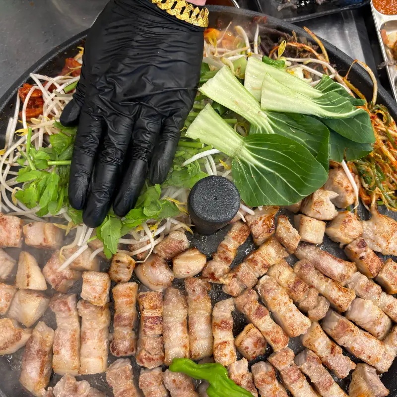 솥뚜껑 위에 삼겹살, 김치, 콩나물, 야채 등이 함께 구워지고 있는 모습