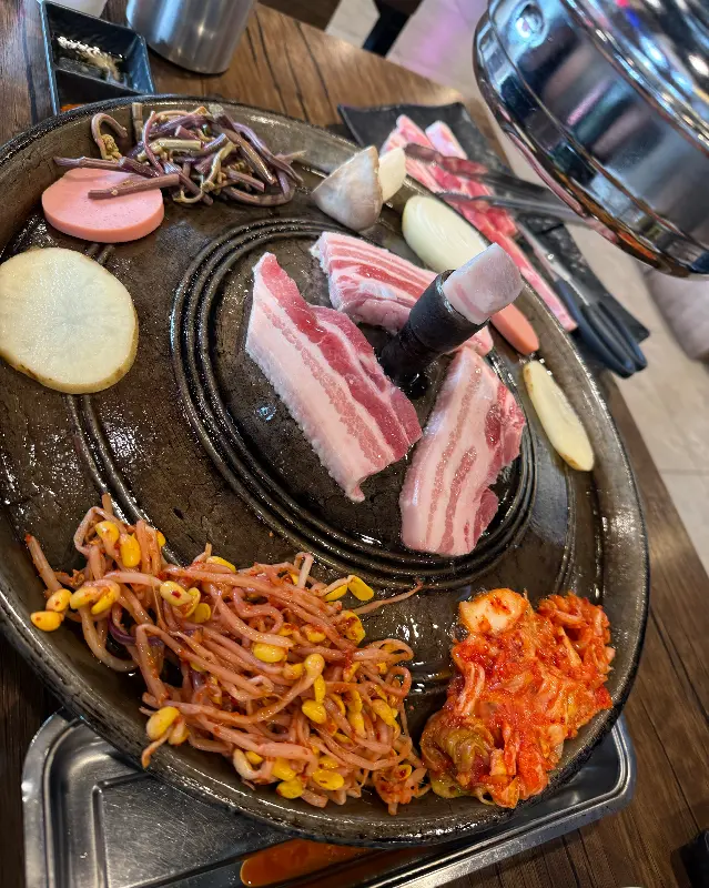 솥뚜껑에 올려진 삼겹살, 김치, 콩나물의 모습