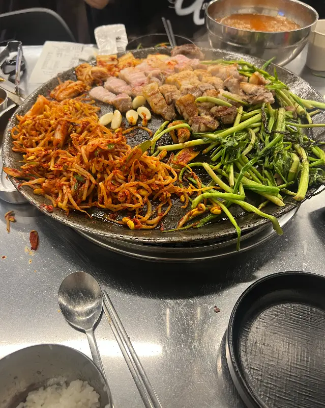 솥뚜껑 위에서 삼겹살, 김치, 콩나물, 미나리가 함께 구워지고 있는 모습