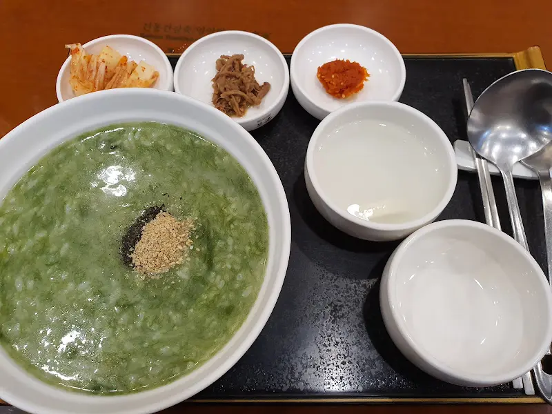 메생이굴죽의 모습, 검은깨와 김 가루가 뿌려져 있고 깍두기, 장조림, 젓갈이 함께 놓여 있다.