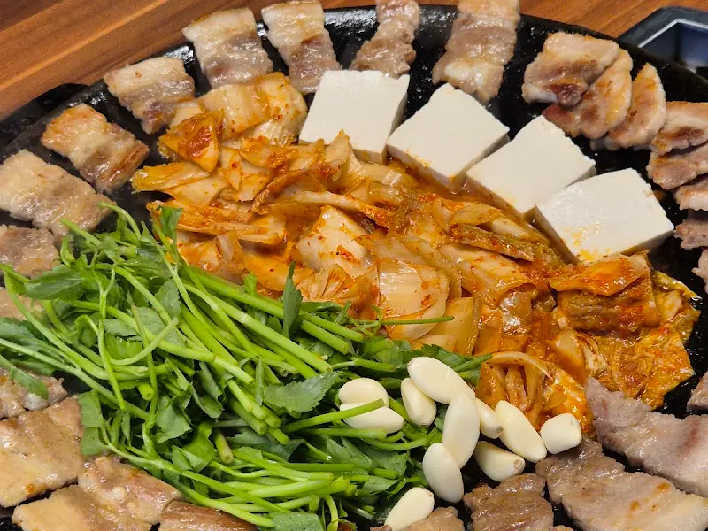 무쇠판에 삼겹살, 김치, 두부, 미나리, 마늘이 함께 구워진 모습