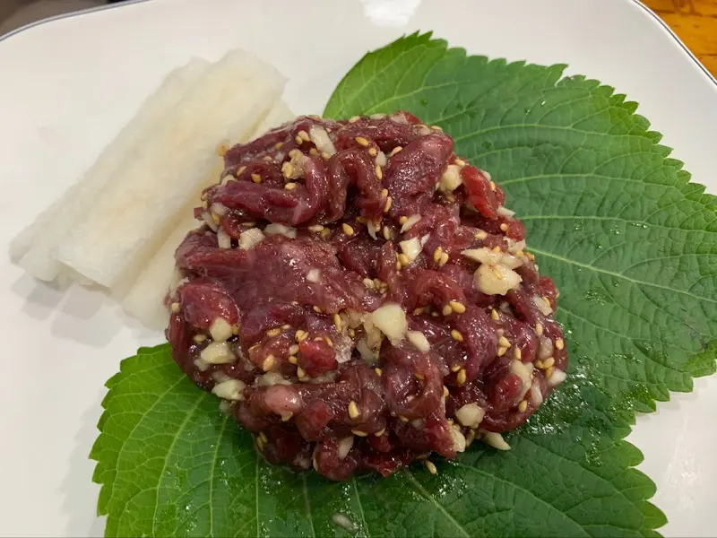 붉은 빛깔의 육회가 싱싱한 깻잎 위에 소담하게 담겨 있다. 옆에는 길게 썰린 배가 가지런히 놓여 있다.