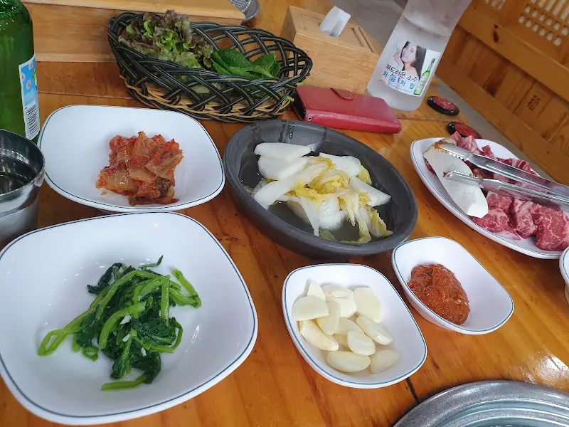 다채로운 밑반찬들이 정갈하게 담겨 나무 테이블 위에 놓여 있다. 백김치, 샐러드, 묵 등 푸짐한 구성이 눈길을 사로잡는다.