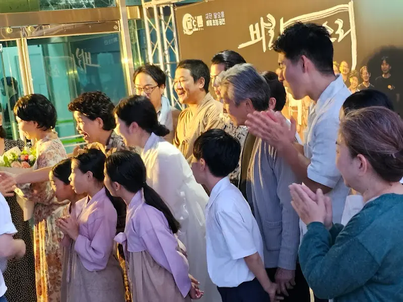 공연이 끝난 후, 배우들이 관객들과 함께 사진을 찍고 있다. 밝은 표정의 배우들과 관객들의 모습이 보기 좋다.