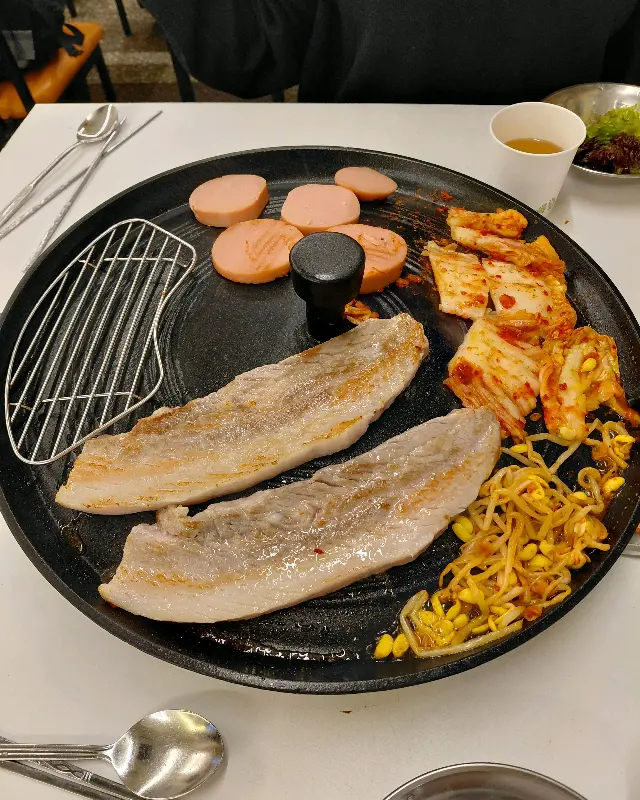 솥뚜껑 위에 삼겹살, 김치, 콩나물, 소세지를 함께 구워 먹는 모습