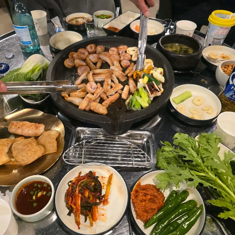 불판 위에 막창과 꽈리고추, 마늘 등이 함께 구워지고 있고, 테이블 위에는 다양한 쌈 채소와 밑반찬들이 놓여있다.