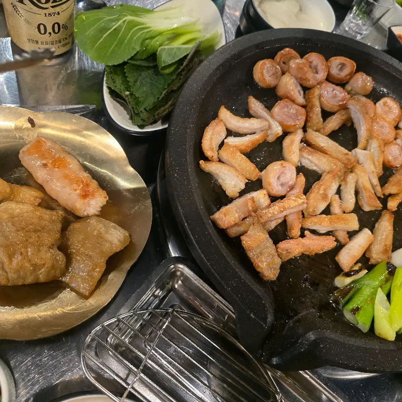 불판 위에 막창과 꽈리고추, 마늘 등이 함께 구워지고 있고, 테이블 위에는 쌈 채소와 소스, 밑반찬 등이 놓여있다.