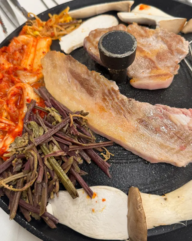 솥뚜껑 위에 삼겹살과 구운 김치, 구운 버섯이 놓여있는 모습