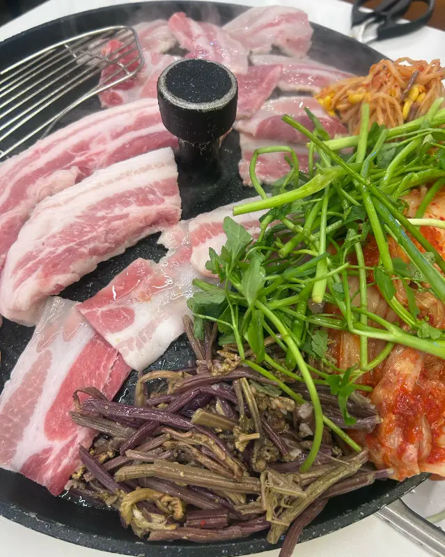 솥뚜껑에 삼겹살, 김치, 콩나물, 고사리, 미나리를 함께 구워 먹는 모습