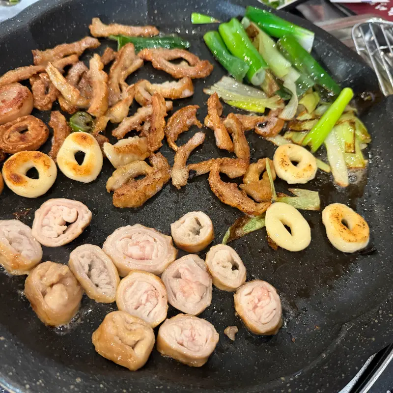 불판 위에서 막창과 꽈리고추, 마늘이 함께 구워지고 있는 모습이 클로즈업 되어 있다.