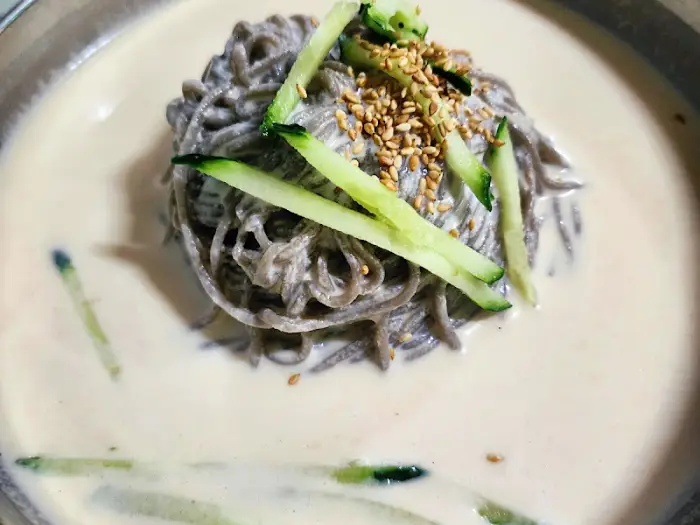 뽀얀 콩국수 국물 위로 얇게 썬 오이와 깨가 올려져 있다.