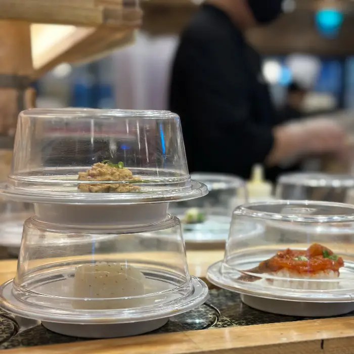 투명한 덮개 아래 놓인 두 개의 초밥. 하나는 하얀 쌀밥 위에 소스가 올라가 있고, 다른 하나는 붉은 양념이 덮여 있다.