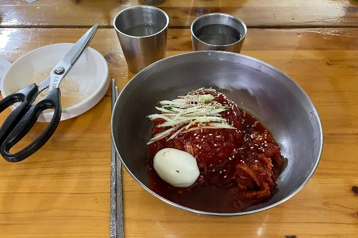 식탁 위에 놓인 비빔밀면과 컵, 젓가락, 가위. 놋그릇의 고급스러움이 느껴집니다.