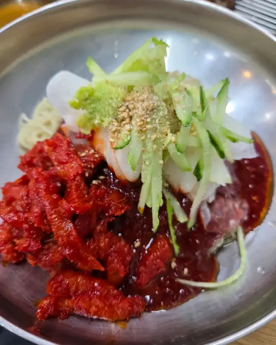 김치와 함께 비벼진 물비빔냉면. 고명으로 오이채, 계란지단, 깨가 뿌려져 있다.