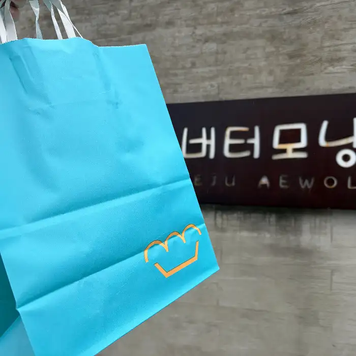 하늘색 쇼핑백에 버터모닝 로고가 그려져 있다. 배경에는 '버터모닝 JEJU AEWOL' 이라고 적힌 검은색 간판이 보인다.