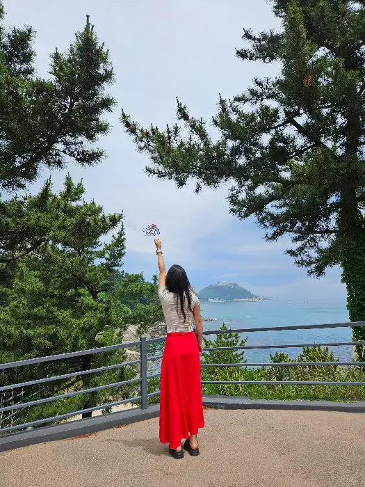 붉은 치마를 입은 여성이 바다를 배경으로 작은 깃발을 들고 서 있다.