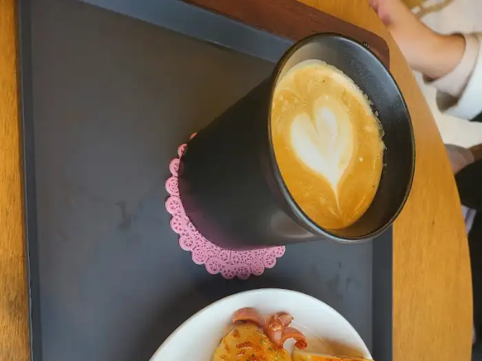 따뜻한 라떼 아트가 그려진 커피와 롱 소시지빵으로 보이는 빵이 함께 담겨 있다.