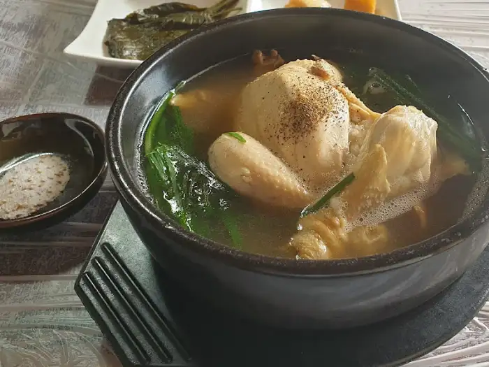 검은색 그릇에 담긴 닭 한 마리가 통째로 들어간 백숙, 곁들임 반찬으로 보이는 작은 종지가 있습니다.