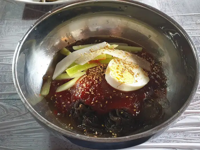 놋그릇에 담긴 비빔냉면, 빨간 양념장 위에 계란 반쪽과 채 썬 오이가 올려져 있습니다.