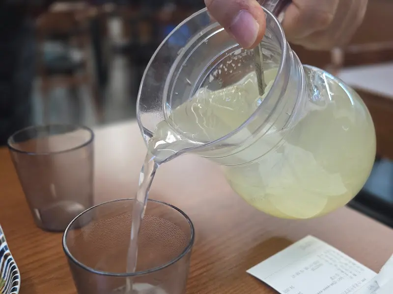 투명한 유리 물통에서 레몬에이드를 컵에 따르는 모습. 얼음이 담긴 컵에 레몬에이드가 쏟아지면서 청량감을 더한다.