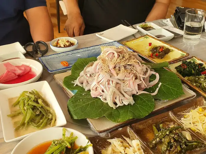 테이블 전체를 채운 다양한 해산물 요리와 밑반찬들. 중앙에는 연한 색의 해산물 샐러드 같은 요리가 놓여 있고, 주변으로 녹색 채소 요리, 젓갈류, 쌈 채소 등이 풍성하게 차려져 있다.