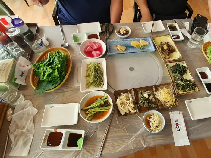 다양한 음식이 놓인 테이블의 전체 모습. 쌈 채소, 여러 가지 밑반찬, 밥, 소스 등이 가지런히 놓여 있으며, 벽면에는 액자와 조명, 에어컨 등이 보인다.