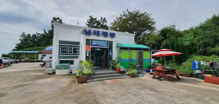 식당 외관 모습. 하얀색 건물에 파란색 간판과 녹색 출입문이 눈에 띈다. 건물 앞에는 테이블과 의자, 그리고 우산이 놓여 있어 야외 공간도 있음을 보여준다.