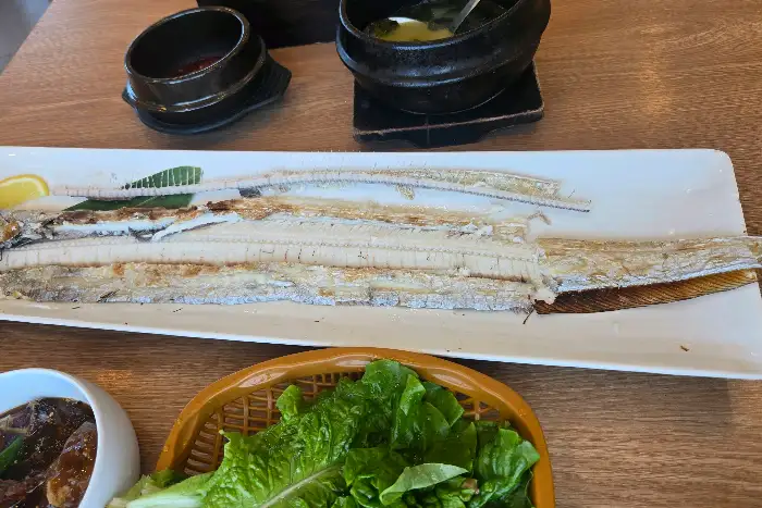 테이블 위에 통갈치구이가 놓여 있고, 쌈 채소가 보인다.