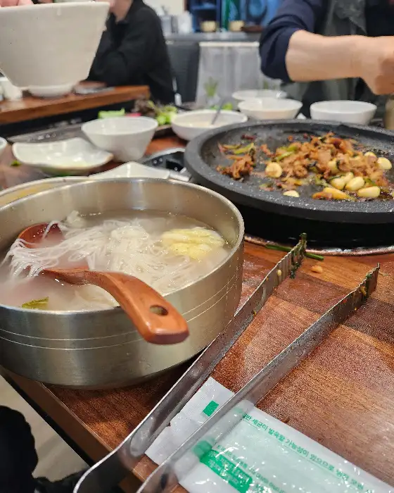 금속 그릇에 담긴 시원한 김치말이 국수, 그 옆에 오리불고기가 조리되고 있는 돌판이 보인다.