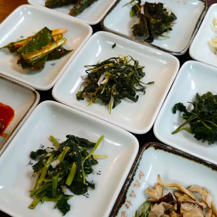 앞서 본 이미지와 비슷한 구성으로, 여러 종류의 나물 반찬들이 놓여 있다. 일부 나물은 양념이 되어 있고, 일부는 본연의 색을 유지하고 있다.