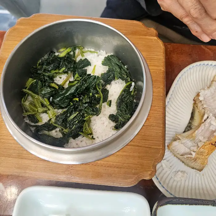 솥밥 그릇의 옆 모습. 밥은 곤드레나물과 함께 섞여 있으며, 밥솥은 나무 받침대에 올려져 있다. 옆에는 밥과 함께 먹을 생선 구이 조각이 접시에 담겨 있다.