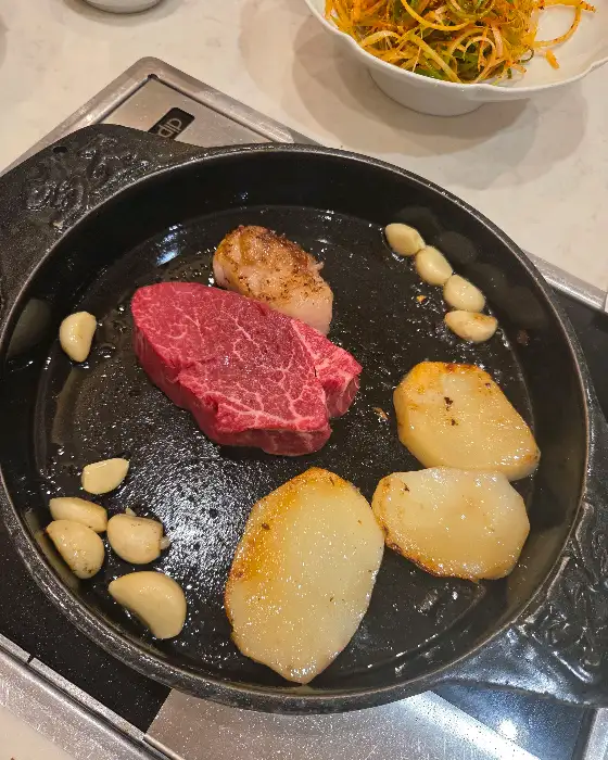 팬 위에는 고기, 감자, 마늘, 그리고 약간의 익힌 관자가 놓여 있다. 배경에는 샐러드와 같은 음식이 담긴 그릇이 보인다.