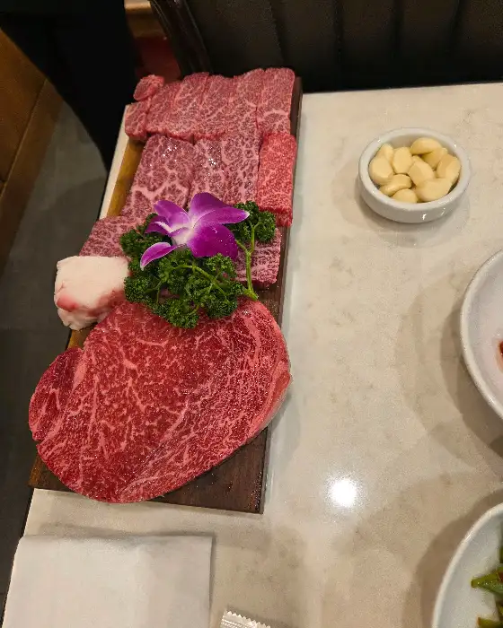 선홍빛 한우 모듬이 나무 도마 위에 화려하게 놓여 있다. 보라색 꽃과 푸른 잎으로 장식되어 있으며, 옆에는 마늘이 담긴 작은 그릇이 보인다.
