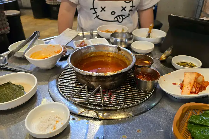 테이블 가운데 연탄불 위에 놓인 숯불 그릴에는 두툼한 흑돼지 오겹살이 구워지고 있습니다. 직원분이 집게로 고기를 뒤집는 모습이 보입니다.