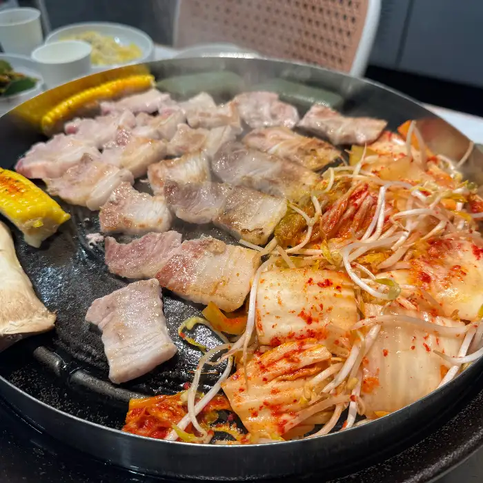 불판에 삼겹살, 김치, 콩나물, 옥수수 등이 함께 구워지고 있습니다.