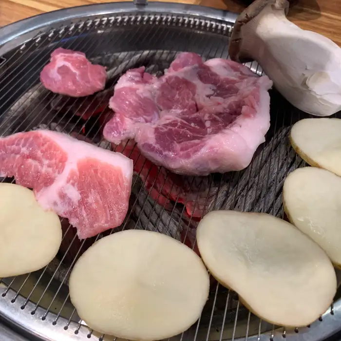 숯불 위에 놓인 신선한 돼지고기 덩어리들과 슬라이스된 버섯, 감자