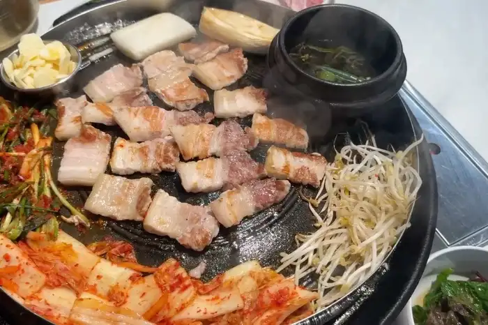 삼겹살과 김치, 콩나물, 버섯 등이 불판 위에서 익어가고 있으며, 찌개가 곁들여져 있습니다.