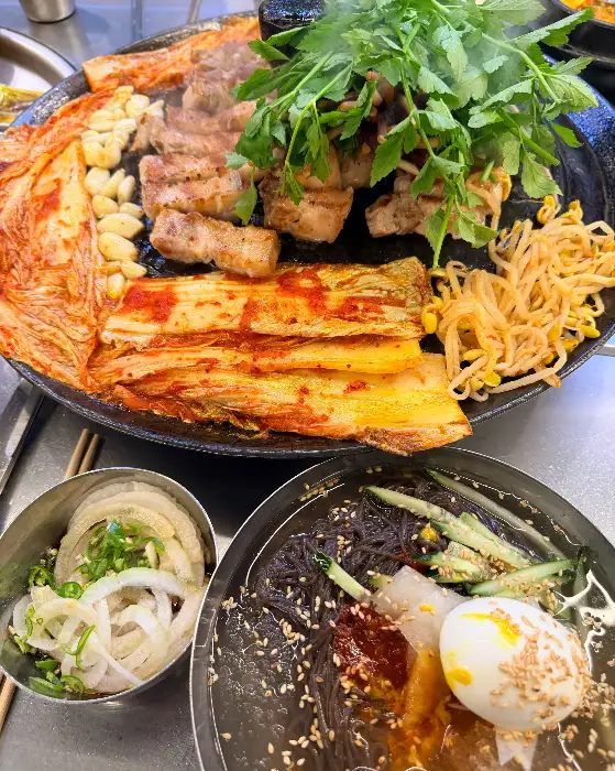 솥뚜껑 위에서 익어가는 고기와 함께 김치, 콩나물, 미나리가 올려져 있다.  전체적인 푸짐함이 느껴진다.