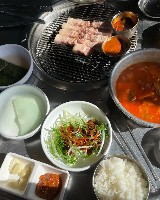 테이블 세팅 전체 모습. 연탄불 위에는 흑돼지 오겹살이 구워지고 있으며, 멜젓과 소스가 담긴 놋그릇이 보입니다. 신선한 파절이, 쌈 채소, 마늘, 밥, 그리고 커다란 뚝배기에는 김치찌개가 담겨 있습니다.