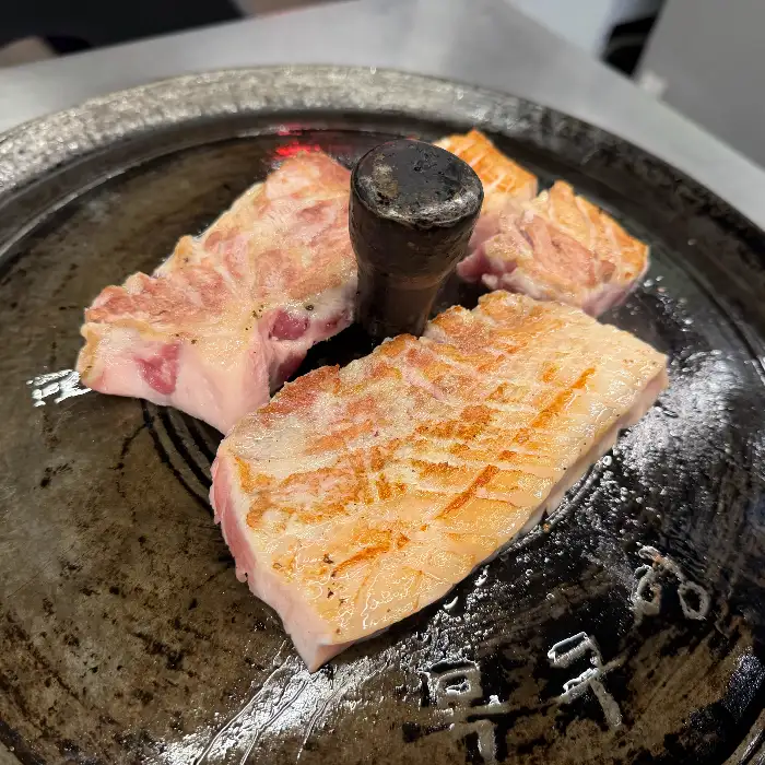 솥뚜껑 중앙의 불꽃 위에 올려진 삼겹살 덩어리.  기름기가 녹아내리며 빛나는 표면이 먹음직스럽다.