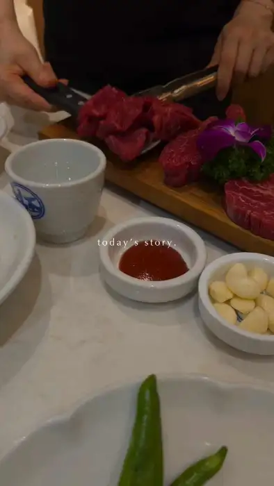 나무 도마 위에 잘라진 고기와 쌈장, 마늘, 그리고 붉은 꽃이 놓여 있다. 셰프의 손이 칼을 들고 있는 모습이 보인다.