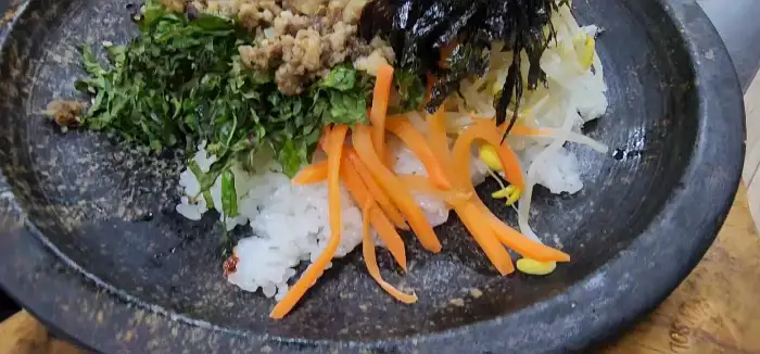 돌솥비빔밥의 클로즈업 사진. 밥 위에 신선한 채소, 다진 고기, 당근채, 김 등이 먹음직스럽게 올라가 있습니다.