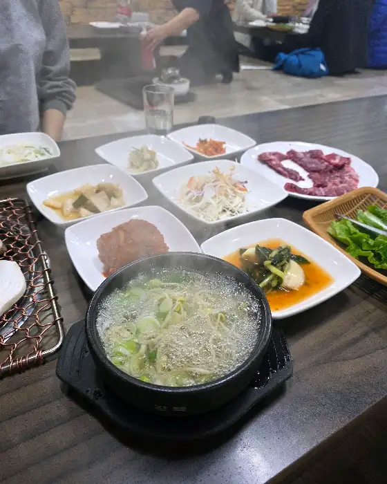테이블 위에 가지런히 놓인 다양한 음식들. 뚝배기 된장찌개, 여러 종류의 밑반찬, 쌈 채소 등이 보입니다.