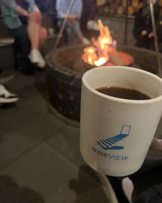캠프파이어 옆에 놓인 나인뷰커피 로고 머그잔