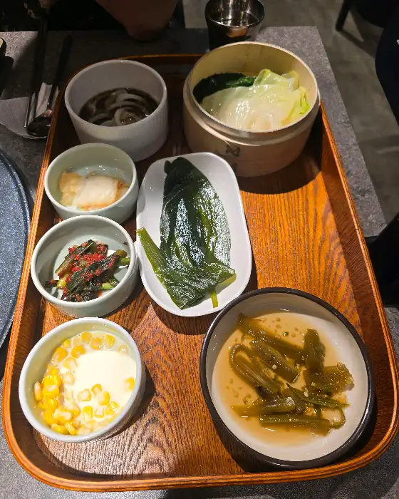 나무 쟁반 위에 다양한 밑반찬이 담긴 여러 개의 작은 그릇들이 놓여 있다. 찐 채소, 김치, 옥수수 콘 등이 보인다.