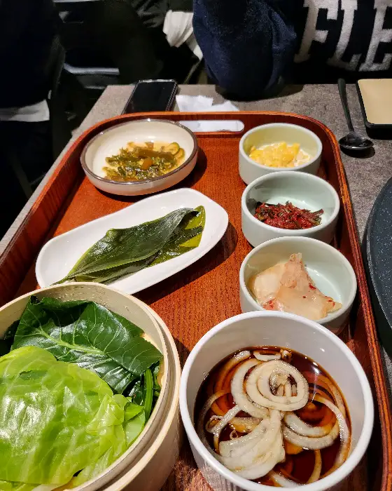 다른 각도에서 촬영된 밑반찬 구성. 찐 채소, 샐러드, 김치 등이 보이며, 쟁반의 나무 질감이 느껴진다.