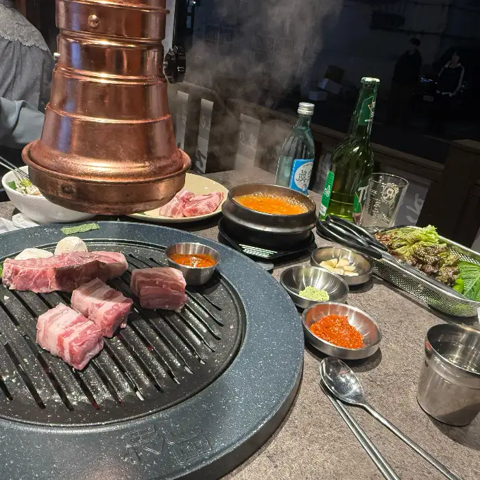 불판 위에서 익어가고 있는 고기와 주변의 밑반찬, 소주병 등이 보이는 테이블 풍경. 숯불 연기가 피어오르고 있다.