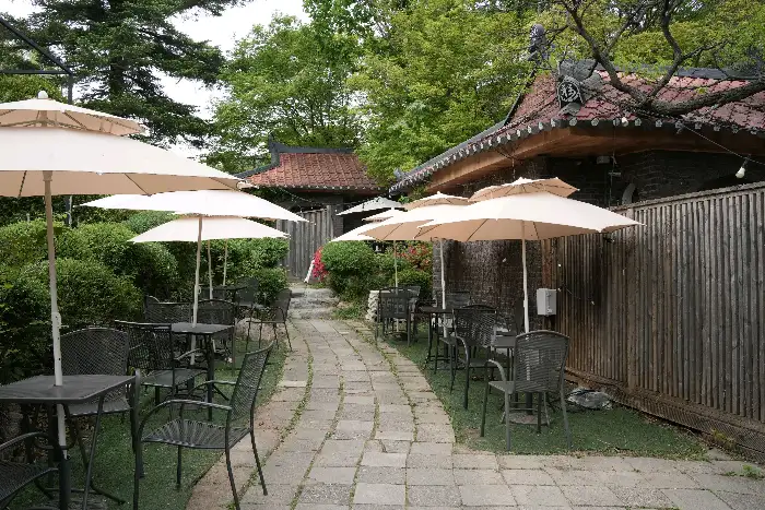 카페 외부에 마련된 야외 테이블과 의자. 우산이 펼쳐져 있고, 나무와 돌이 어우러진 자연 친화적인 모습이다.