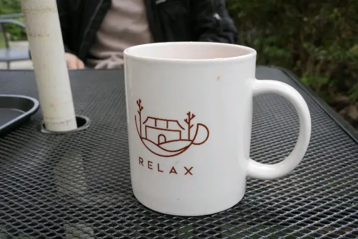 RELAX 로고가 새겨진 흰색 머그잔. 테이블 위에서 커피잔이 놓여 있다.